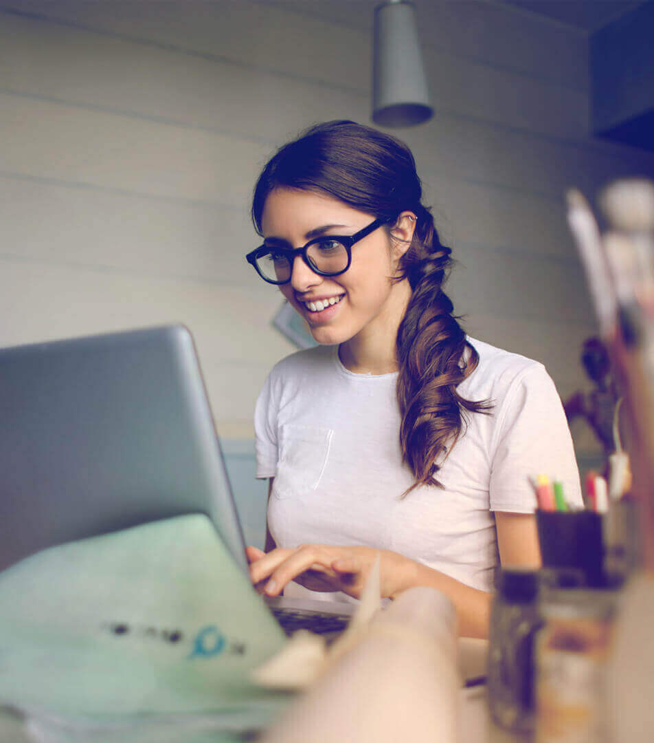 A girl with glasses and brown hair in a ponytail sitting at a desk while smiling and working on her laptop.